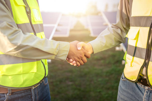 two asian male engineer wearing safety vest handshake with solar panels background. business project deal and agreement in industrial of renewable and green energy concept.